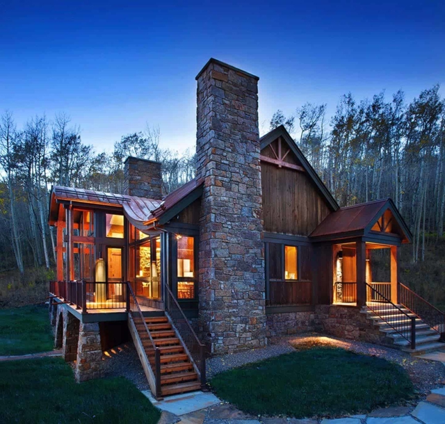 A wood and stone cabin with tall chimneys. The photo is taken during blue hour so the yellow light from the inside provides a warm and inviting contrast to the surrounding. The cabin is surrounded by a forest of tall birch trees.